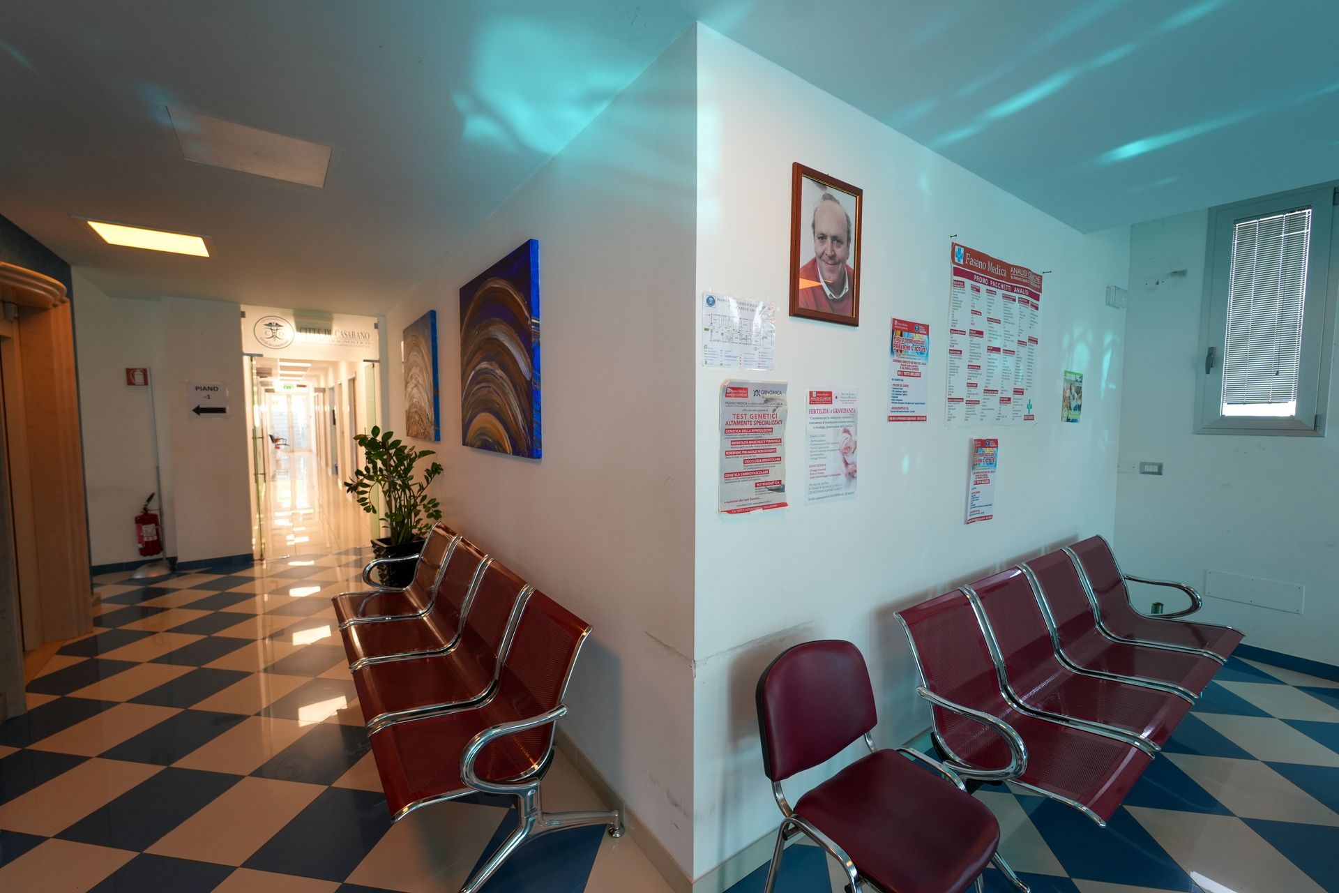 Corridoio di uno studio medico con sedie rosse, poster e pavimento a scacchi.