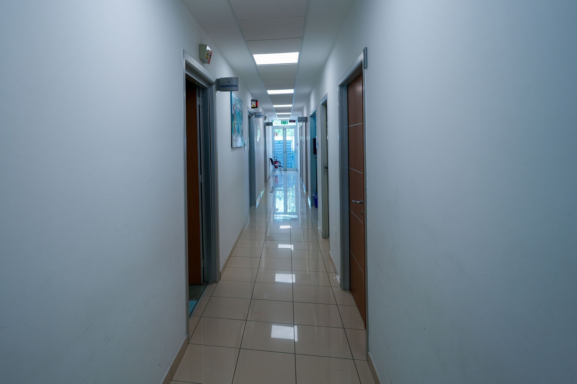 Lungo corridoio con porte su entrambi i lati, illuminato da luci soffuse e una finestra in fondo.