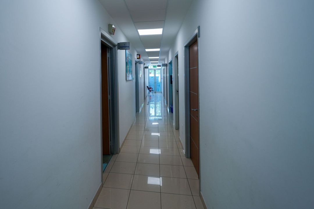 Lungo corridoio con porte su entrambi i lati, illuminato da luci soffuse e una finestra in fondo.