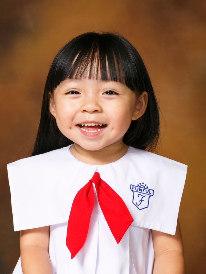 棕色背景下，身穿白色制服、繫著紅色領帶、黑髮的年輕女孩微笑著。