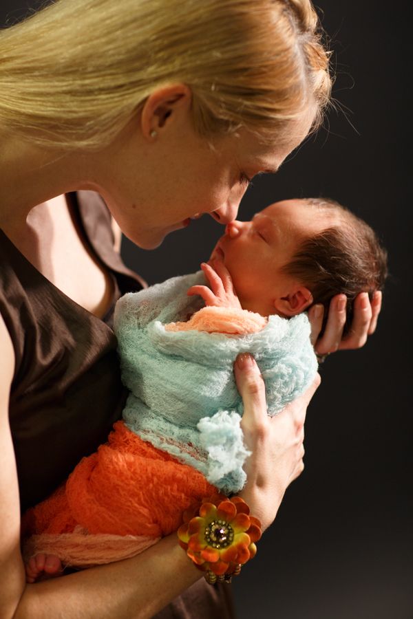 一位金髮女子抱著新生兒，鼻子貼著鼻子。嬰兒裹著藍橙色的布。背景昏暗。