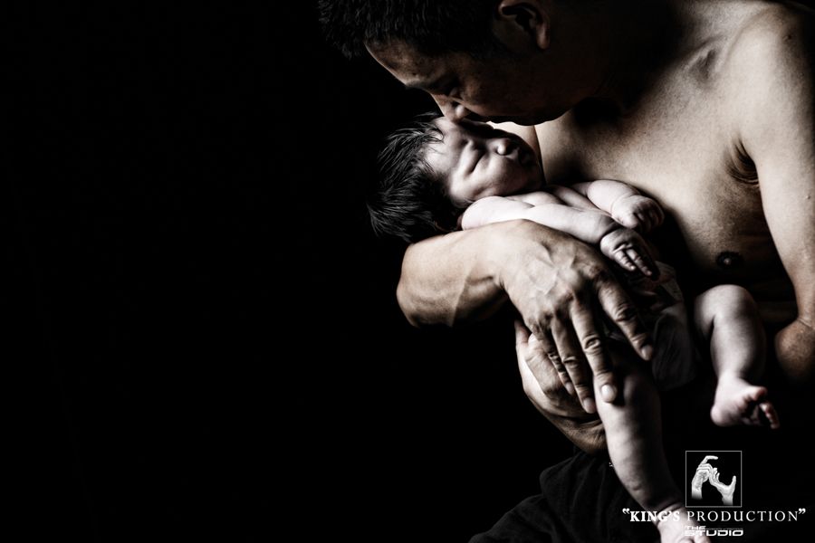 父親親吻新生嬰兒的額頭；兩人都有黑髮，背景為黑色。