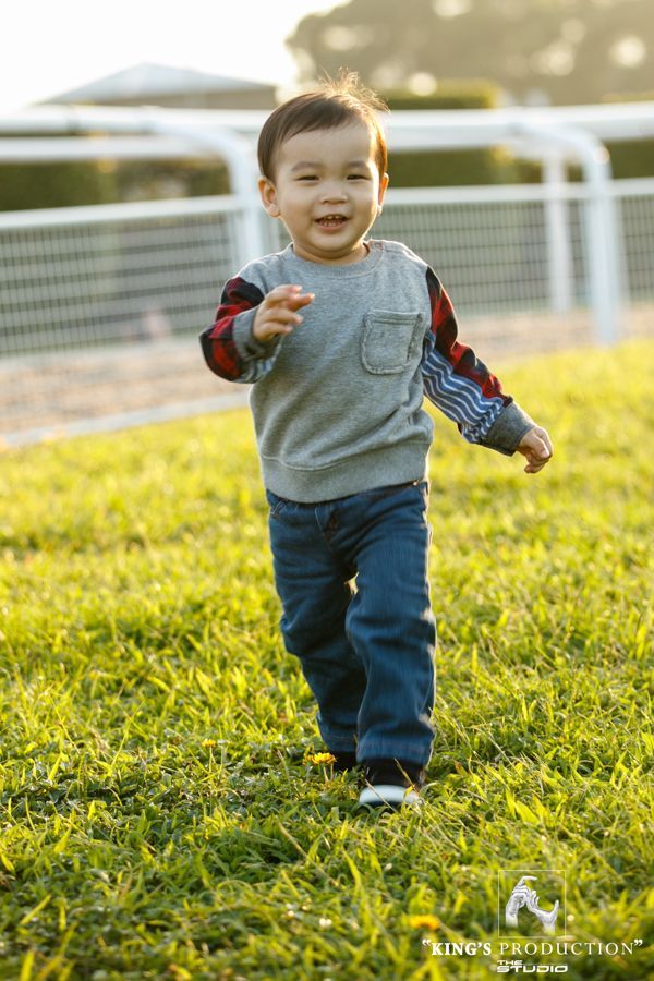 一個微笑的幼兒穿著牛仔褲和灰色運動衫在草地上奔跑。