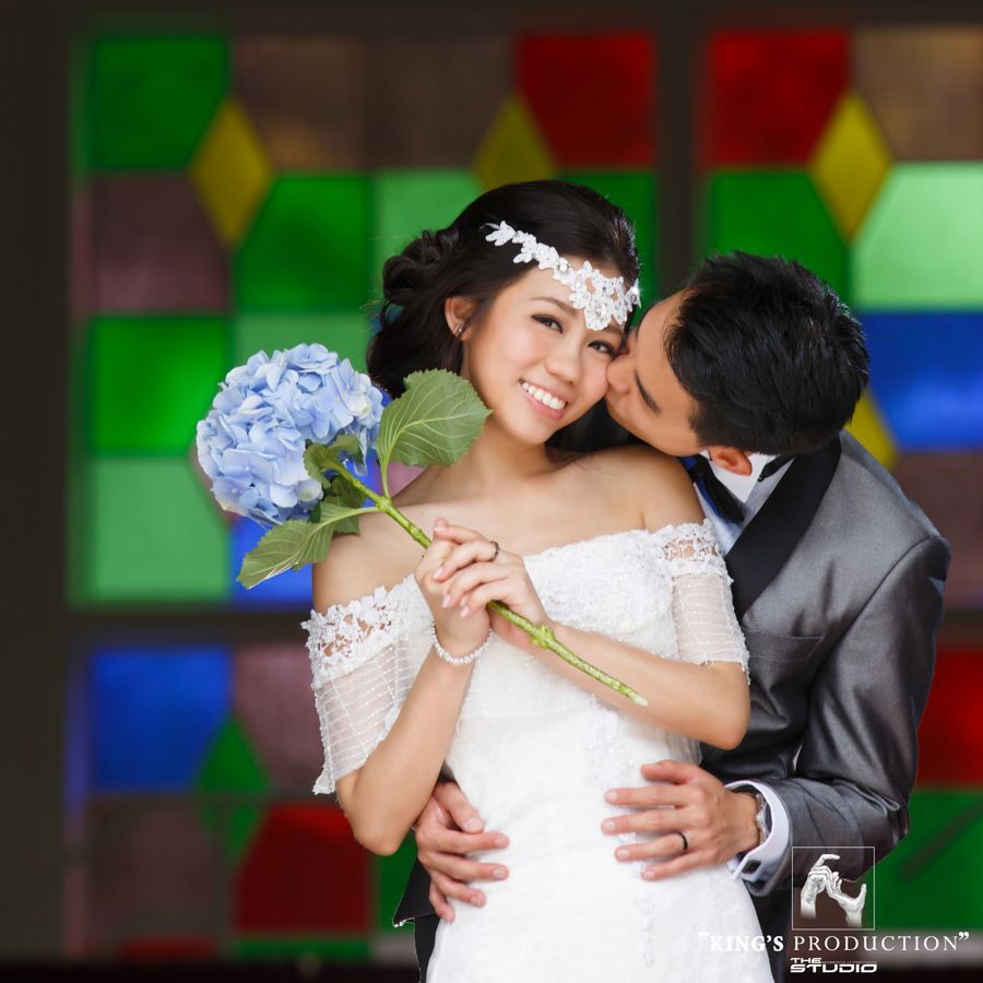 新郎新娘擁抱在一起，新娘手捧藍色繡球花，新郎親吻她的臉頰。背景是彩色玻璃。