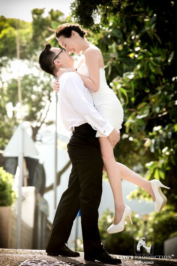 身穿黑褲白襯衫的新郎抱起身穿白裙的新娘。兩人在戶外樹葉旁微笑。