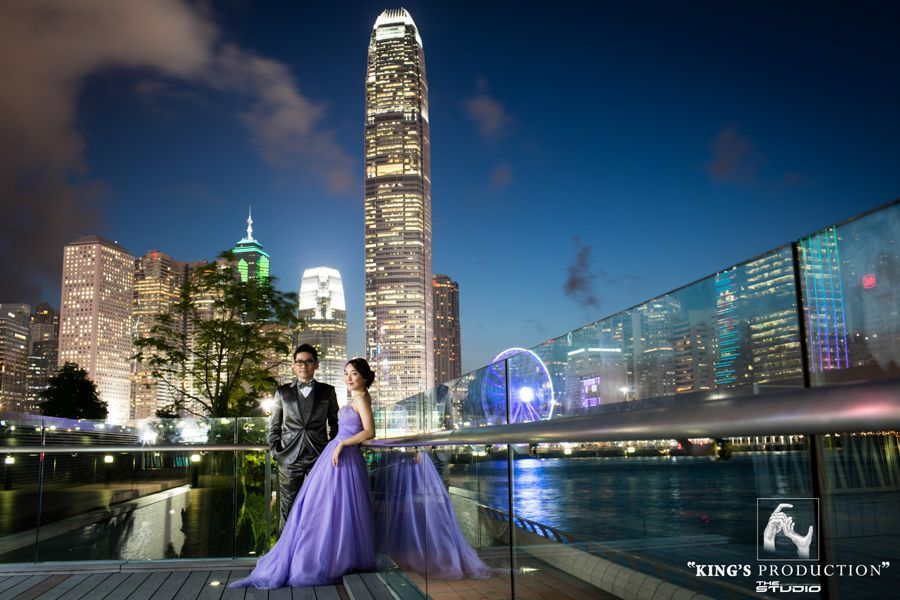 夜晚，一對穿著正式服裝的夫婦站在海濱平台上，欣賞城市天際線，包括一座高大的燈光建築和摩天輪。