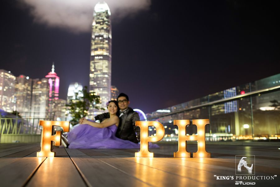 一對情侶在夜晚的城市天際線下，在發光的“PH”字母前擺姿勢拍照。女子身著紫色連身裙，男子身穿黑色夾克。