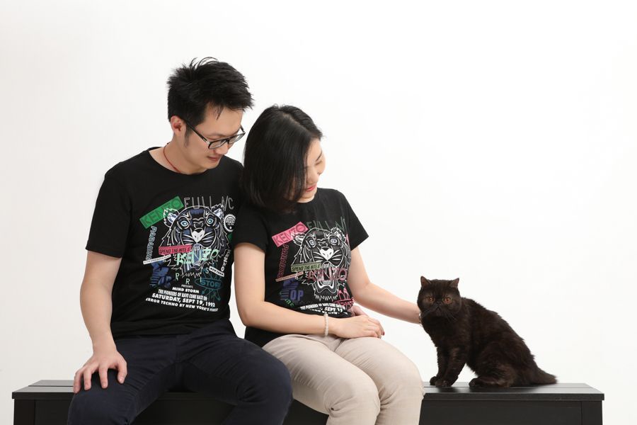 一對穿著黑色 T 恤的夫婦和坐在長凳上的黑貓；白色背景。