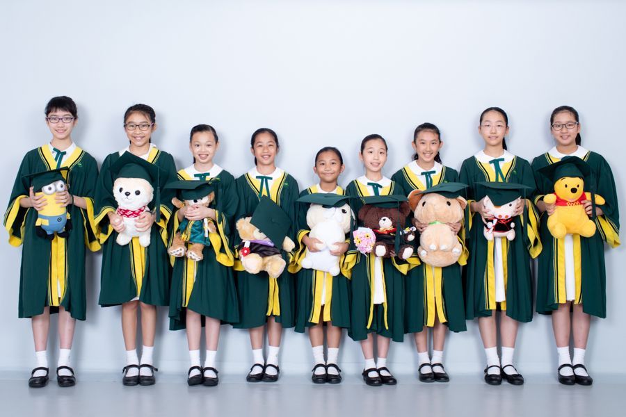 十名身穿畢業禮服的學生手裡拿著毛絨玩具，在灰色的牆壁前微笑。