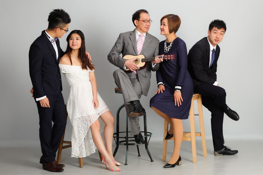 一家人穿著正式服裝，面帶微笑。一位男士坐著彈奏尤克里裡琴，其他人則倚靠在旁邊。背景為灰色。