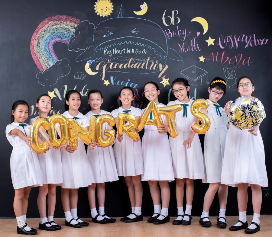 一群身穿白色洋裝的女孩手捧寫有「CONGRATS」的金色氣球，站在黑板前慶祝畢業。