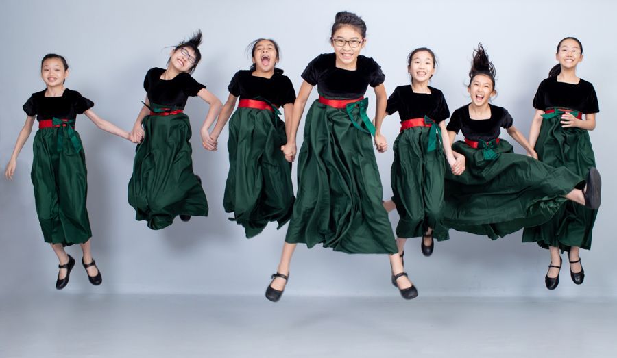 一群穿著綠色洋裝的女孩跳躍著，長髮飄揚。她們在灰色的背景下微笑著，大笑著。