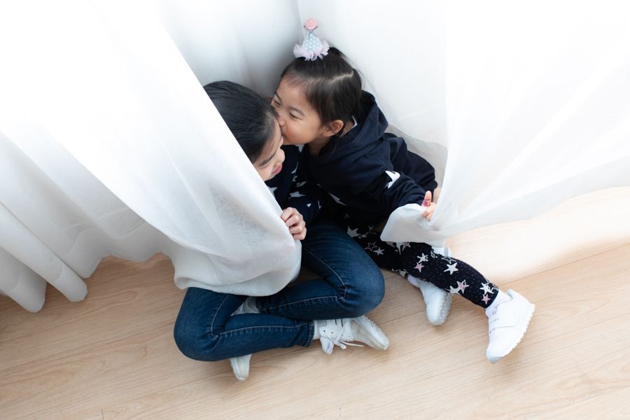 兩個穿著休閒服裝的年輕女孩在白色窗簾後面擁抱，一個親吻另一個的臉頰。