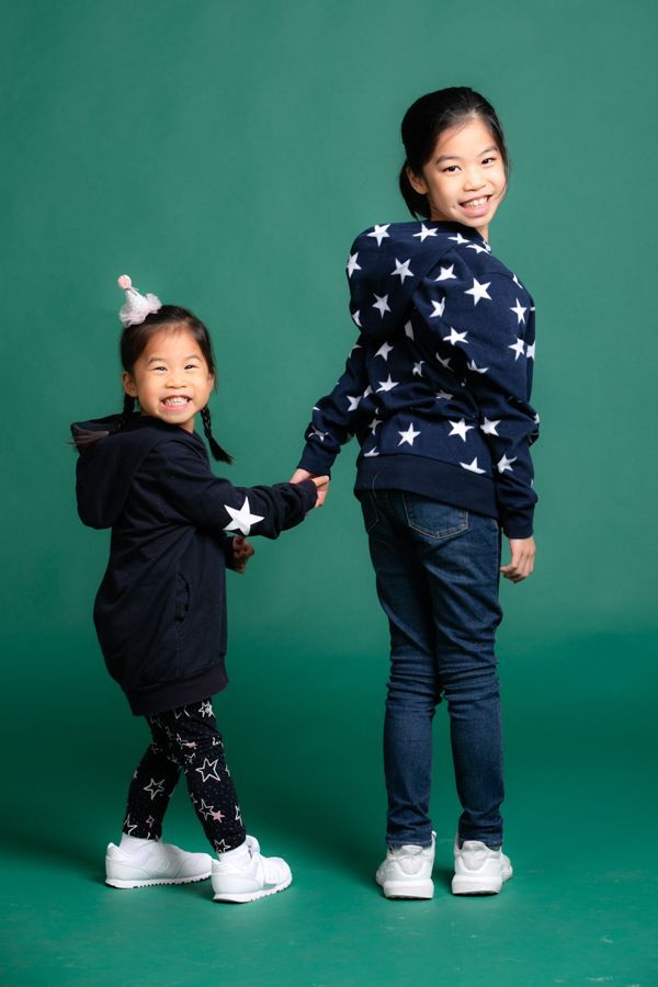 兩個小女孩手牽手，面帶微笑。一個穿著星星圖案的連帽衫，另一個戴著派對帽。背景為綠色。