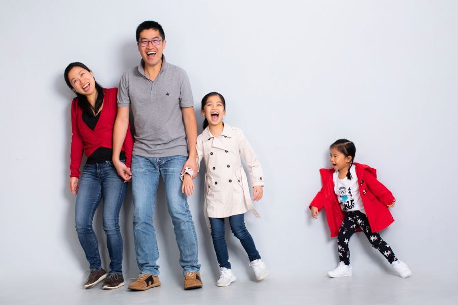 一家五口倚著灰色的牆，面帶微笑，嬉笑不止。兩個女孩穿著夾克，其中一個還在動。