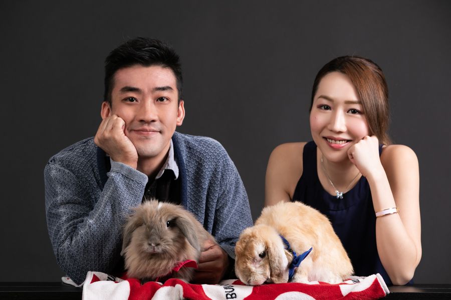 一對面帶著微笑的夫婦在黑色背景上與兩隻寵物兔子合照。