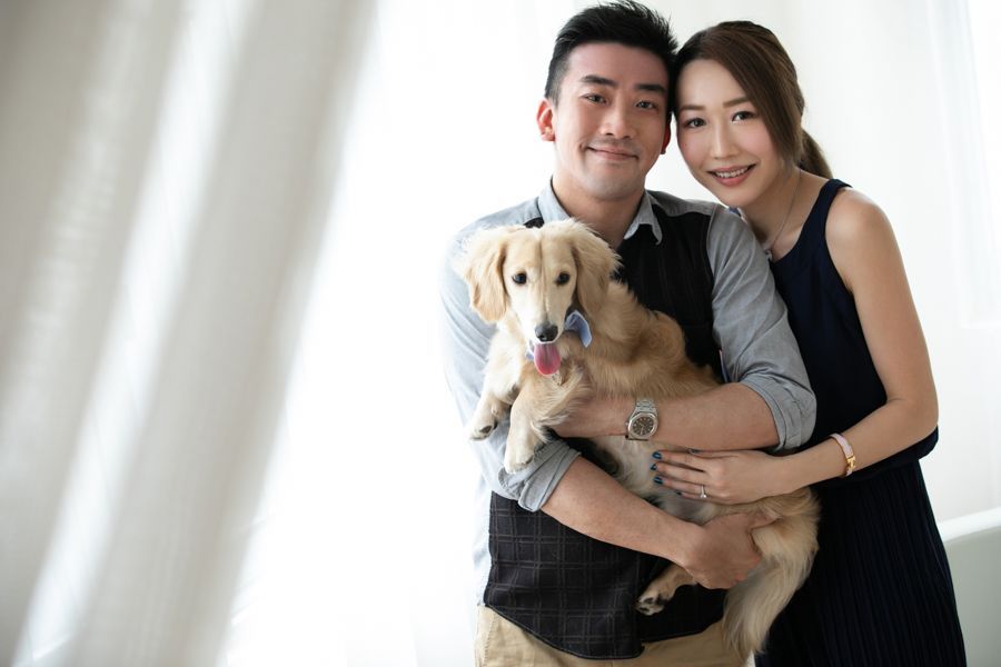一對微笑的夫婦在室內抱著一隻金毛狗，背景是明亮的白色背景。