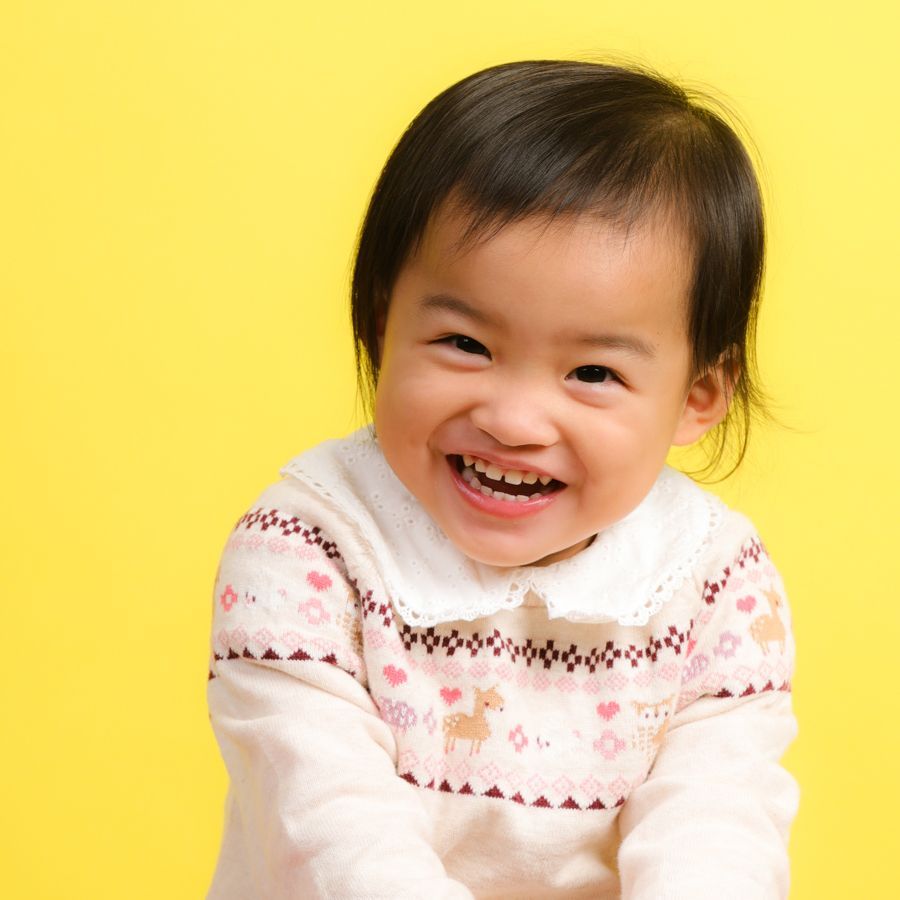 在明亮的黃色背景下，一頭黑髮、身穿圖案的毛衣的幼兒微笑著。