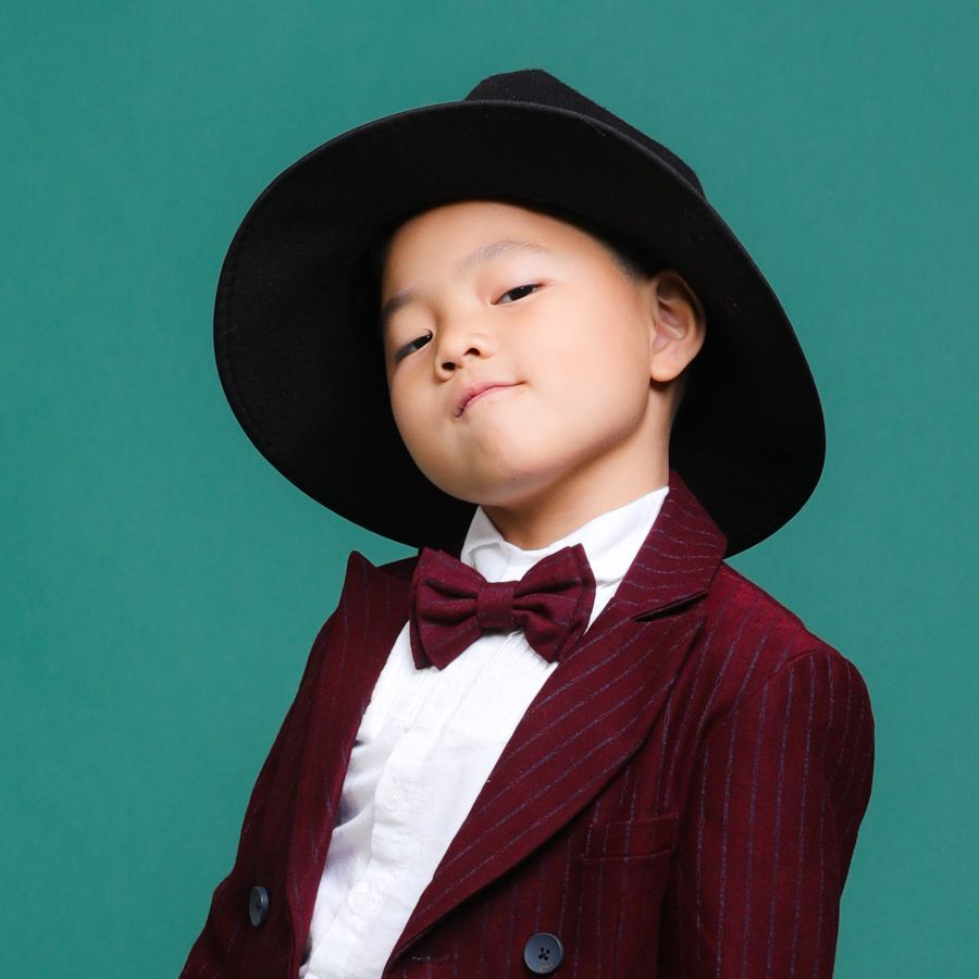身穿酒紅色西裝、繫著領結、戴著黑色帽子的亞洲小孩在綠色背景下假笑。