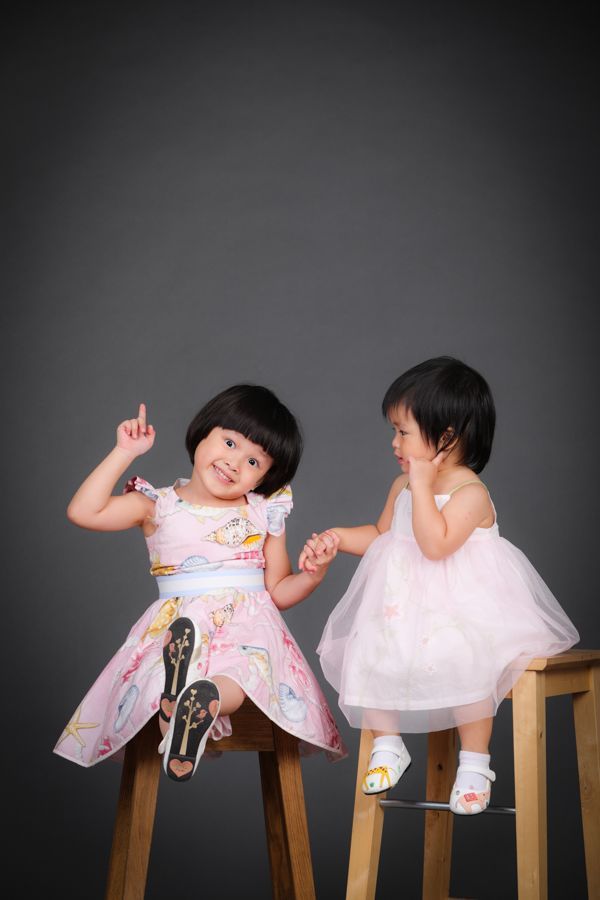 兩個年輕女孩，面帶微笑，手牽手。其中一個女孩面朝上，身穿粉紅色連身裙，坐在凳子上，背景是灰色的。