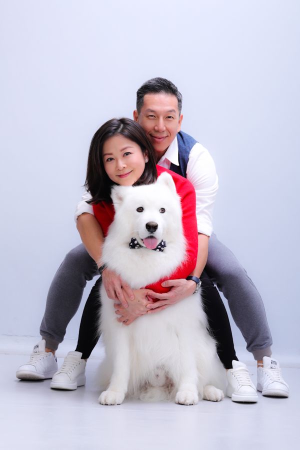 一對夫婦抱著一隻毛茸茸的大白狗，都穿著休閒的衣服，在淺灰色的背景下擺姿勢。