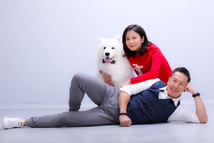 一對夫婦與一隻毛茸茸的白狗合影。女士身穿紅色毛衣，男士身穿背心，兩人都面帶微笑。白色背景。