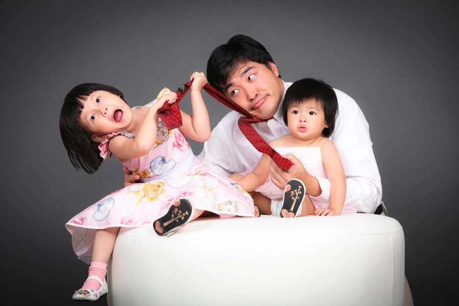 灰色背景下，白色墊子上，父親和兩個小孩嬉戲地拉著領帶。