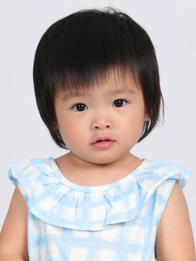 一個有著深色頭髮和瀏海的小孩，穿著藍白相間的連身裙，直視觀眾。