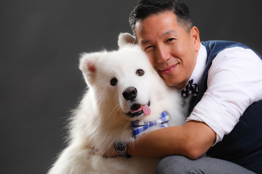 一名男子抱著一隻戴著藍色領結的毛茸茸的白狗；兩人都在灰色背景上微笑。