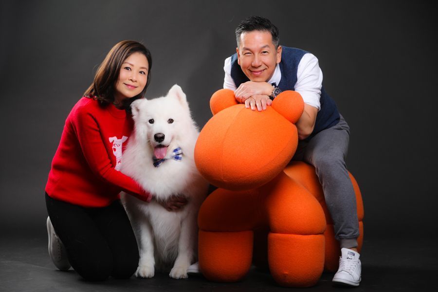 灰色背景上，一男一女，帶著一隻毛茸茸的白狗和一座橘色雕塑。男人倚在雕塑上休息。