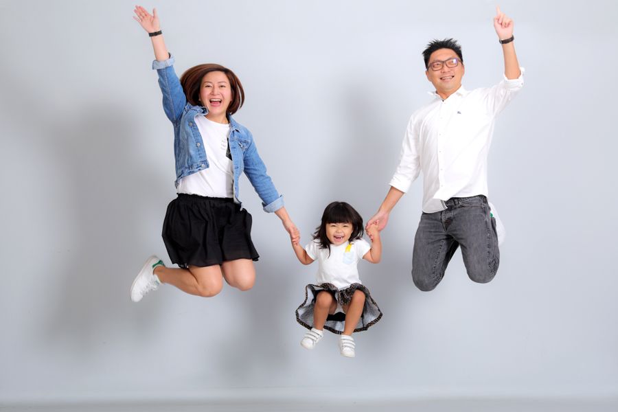 一家三口在灰色背景下跳起，微笑著，手牽手。