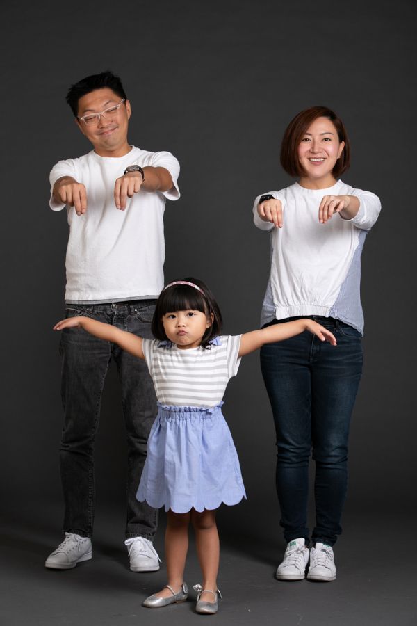 一家三口在灰色背景上擺出張開雙臂、指向下方的姿勢。