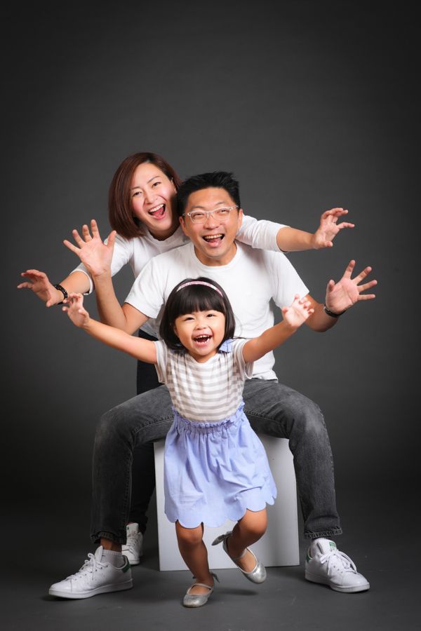 一家三口，笑容滿面，張開雙臂擺姿勢。灰色背景，白色和藍色的衣服。