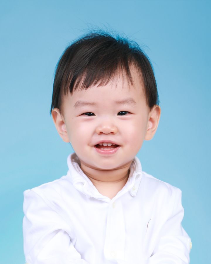 藍色背景中，一頭黑髮、穿白襯衫、面帶微笑的幼兒。