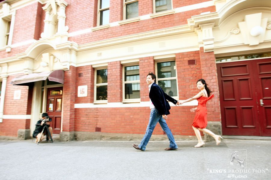 一對情侶，穿著紅色衣服，手牽手，在磚造建築旁散步，旁邊還有攝影師。