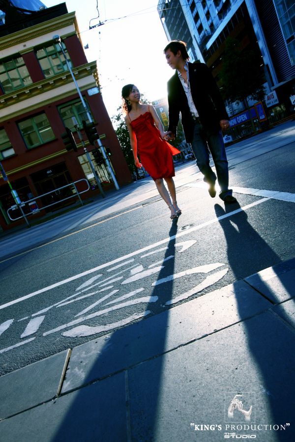一對情侶手牽手，穿過城市建築前的街道。穿著紅裙的女子。長長的影子。