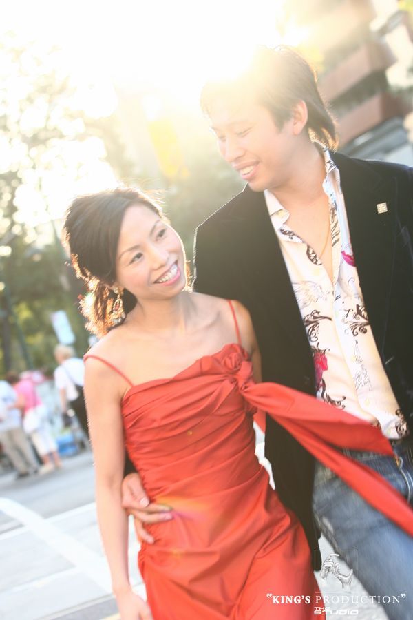 一對情侶邊走邊笑。女子身穿紅色連身裙，男子身穿黑色夾克和花襯衫；陽光明媚。