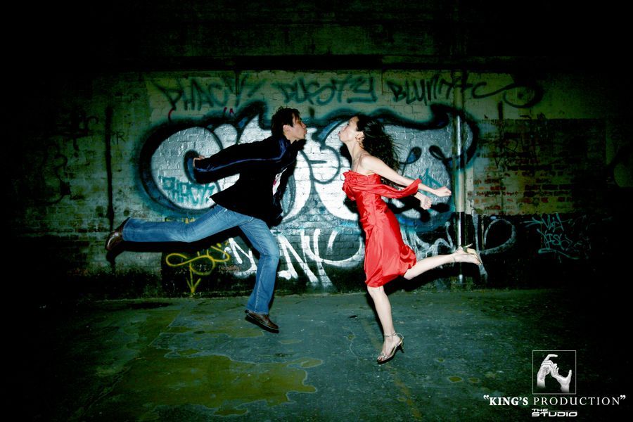 一對情侶在塗鴉牆前跳躍接吻。男士身穿夾克/牛仔褲，女士身穿紅裙。城市場景，氣氛憂鬱。