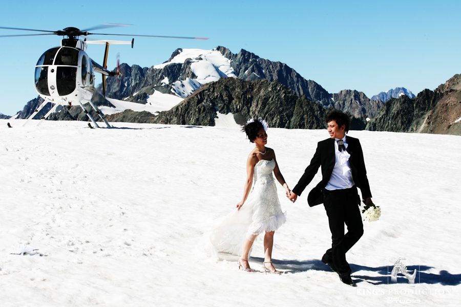 一對新婚夫婦在雪地上走向一架直升機，背景是山脈。陽光明媚。
