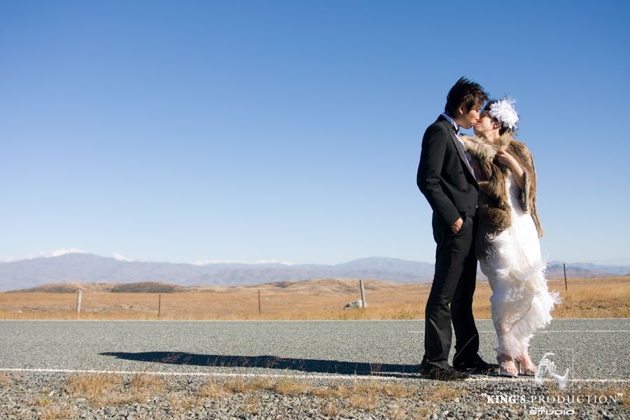 一對情侶在路上接吻，遠處群山環繞，天空湛藍。新娘身穿皮草大衣，新郎穿著西裝。