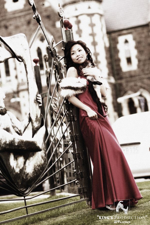 身穿紅色洋裝的女子倚靠在一棟磚造建築前的金屬雕塑上。