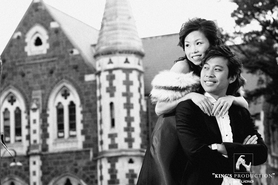 一對身著正裝的情侶在一座石頭建築前擁抱。女子微笑，雙手搭在男子肩上。黑白照片。