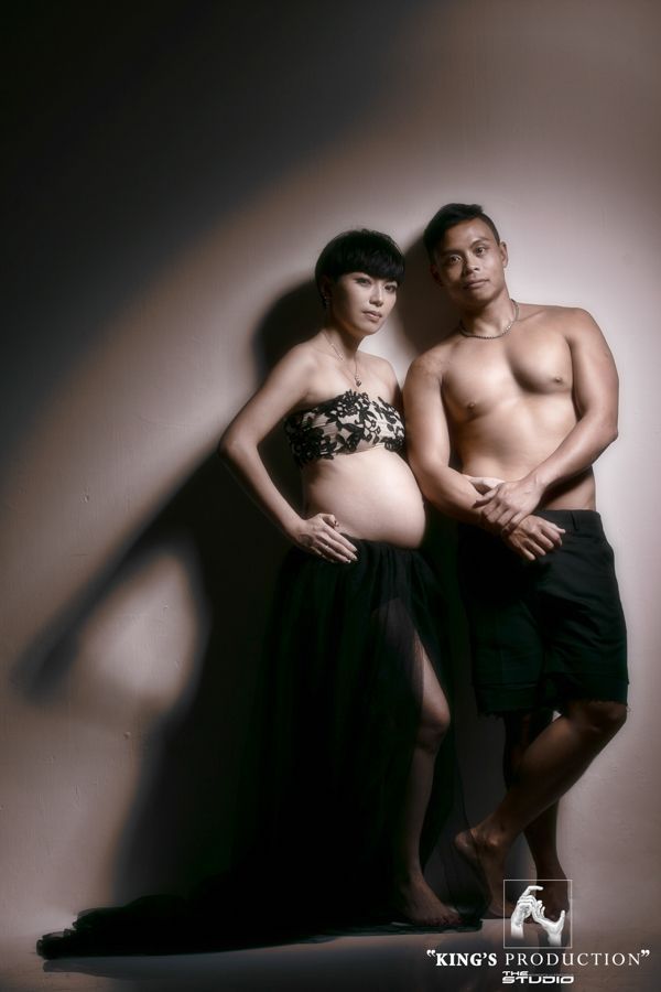孕婦和赤裸上身的男子一起擺姿勢，女人穿著露肩上衣和飄逸的裙子，男人穿著短褲，攝影棚燈光。
