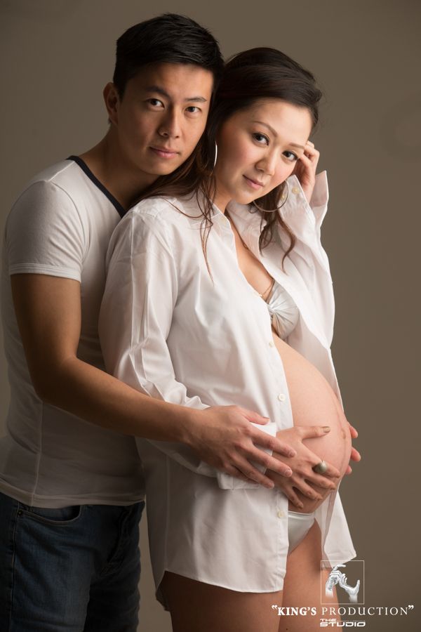 一對情侶正在拍攝孕婦照；男士手臂環繞著女士的腹部。白襯衫，牛仔褲，中性背景。