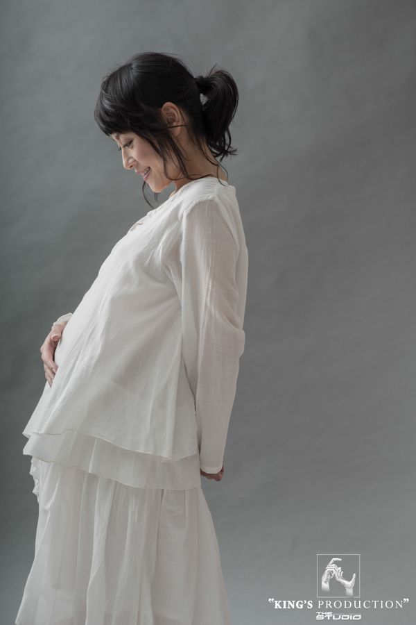 身穿白色服裝的孕婦，微笑著，手放在腹部，背景為灰色。