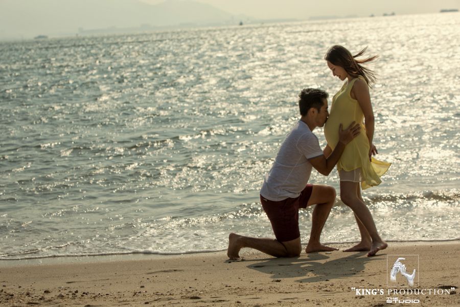 陽光明媚的海灘上，一名男子跪下，親吻孕婦的肚子。