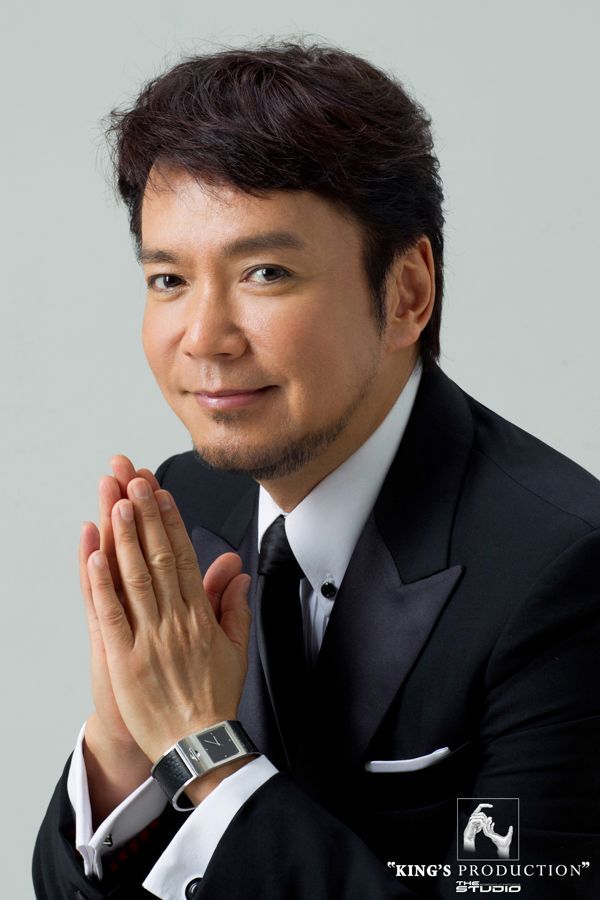 男人身穿黑色西裝，雙手緊握，面帶微笑。