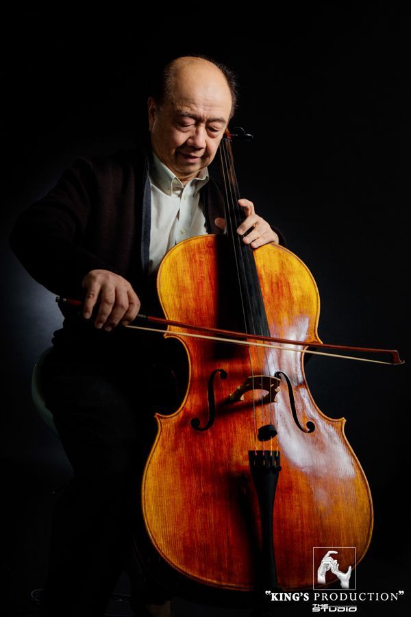 一名男子在黑暗的背景中拉大提琴。他手持琴弓，身穿棕色大提琴，穿著正式服裝，表情專注。