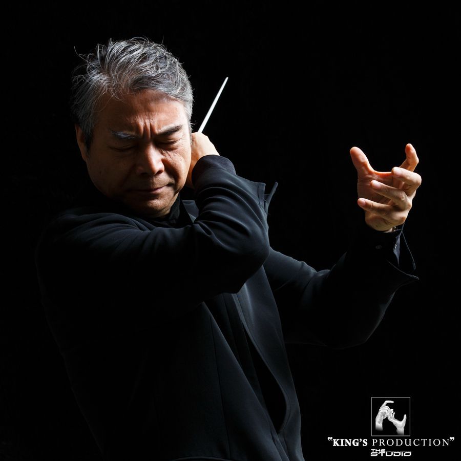 指揮家頭髮花白，穿著黑色服裝，手持指揮棒，在黑暗的背景下表情專注地指揮著。