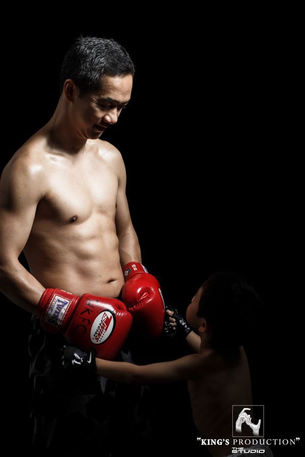 父親赤裸上身，戴著紅色拳擊手套，低頭看著伸手拿手套的孩子。背景昏暗。